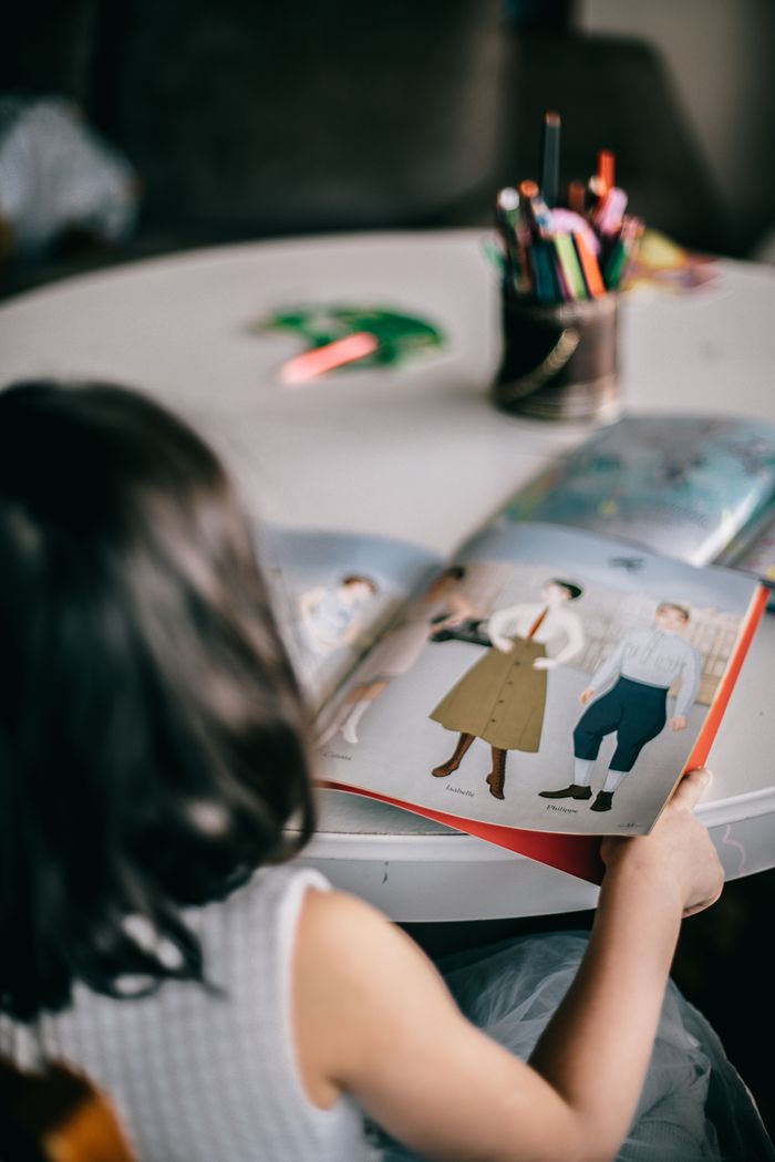 Back view of unrecognizable little girl sitting at table and turning pages of book with pictures